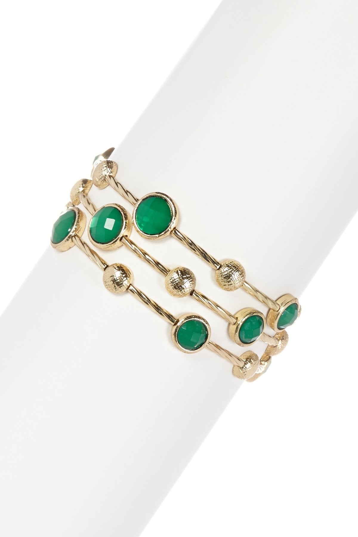 The zirconmania Bracelet Multi-Wire Gold and Beads Bangle 22B0241A features three overlapping gold bands with round green stones and textured gold discs, displayed on a white cylinder—an elegant accessory for any occasion.