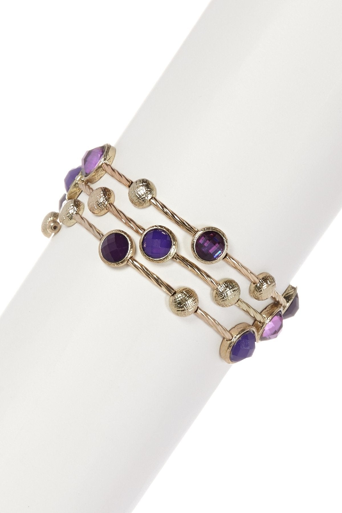 The zirconmania Bracelet Multi-Wire Gold and Beads Bangle 22B0241A features three gold-tone strands with faceted round purple gemstones and textured gold beads, shown on a white cylindrical stand.