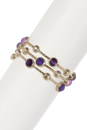 The zirconmania Bracelet Multi-Wire Gold and Beads Bangle 22B0241A features three gold-tone strands with faceted round purple gemstones and textured gold beads, shown on a white cylindrical stand.